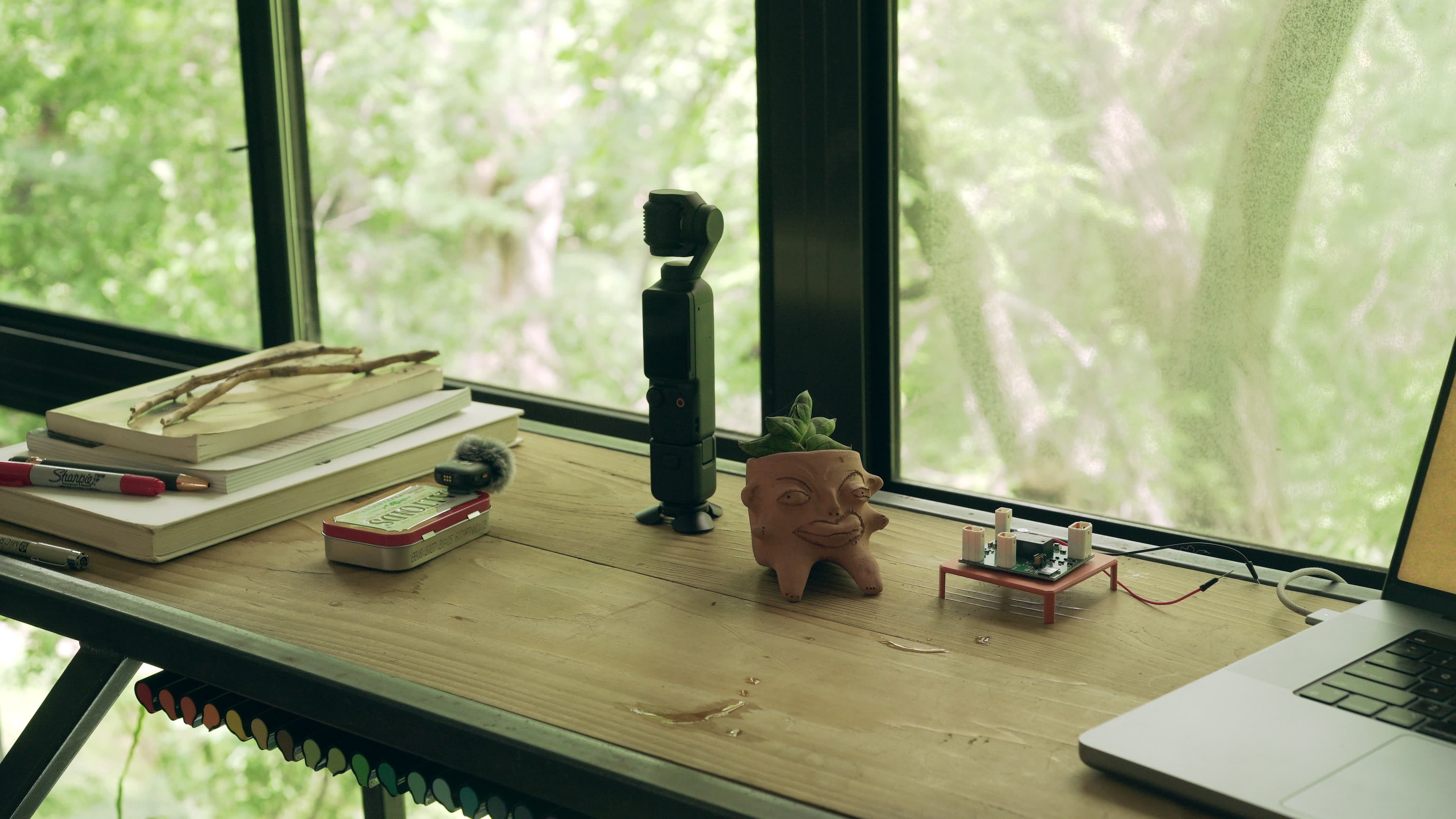 The Gentle Suggestions workbench in Wimberley, Texas  -  notebooks, an Altoids tin of parts, a camera, a clay planter named Monroe, and a circuit board on a small red stand, in front of a window full of trees.